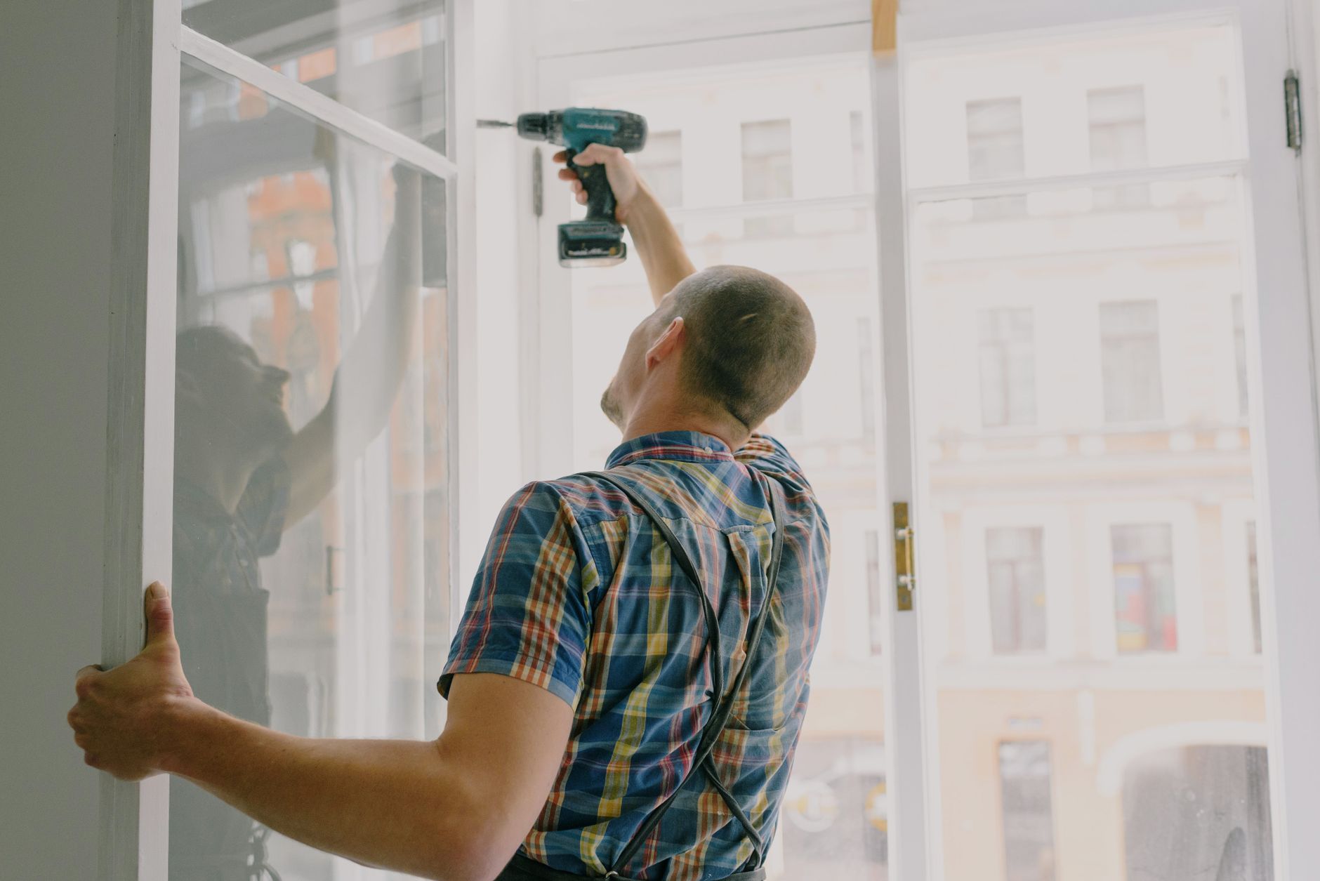 Back view of worker in checkered shirt drilling window frame during renovation process in apartment in Riverside, CA