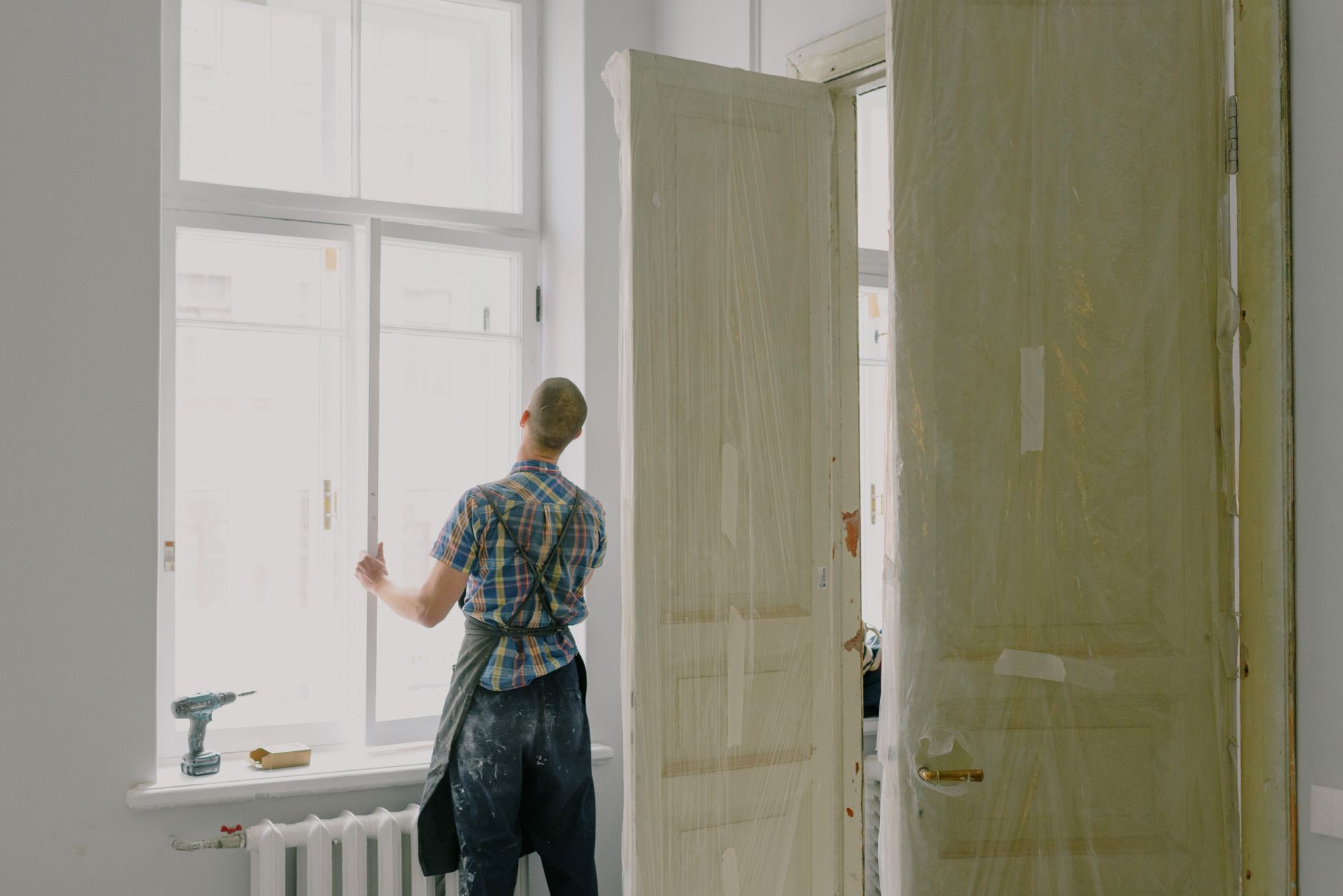 Back view of unrecognizable workman in apron mounting window near drilling machine and doors in flat in Riverside, CA
